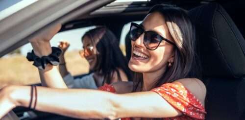 Two young female friends laughing and singing while driving a car on road trip on beautiful summer day.